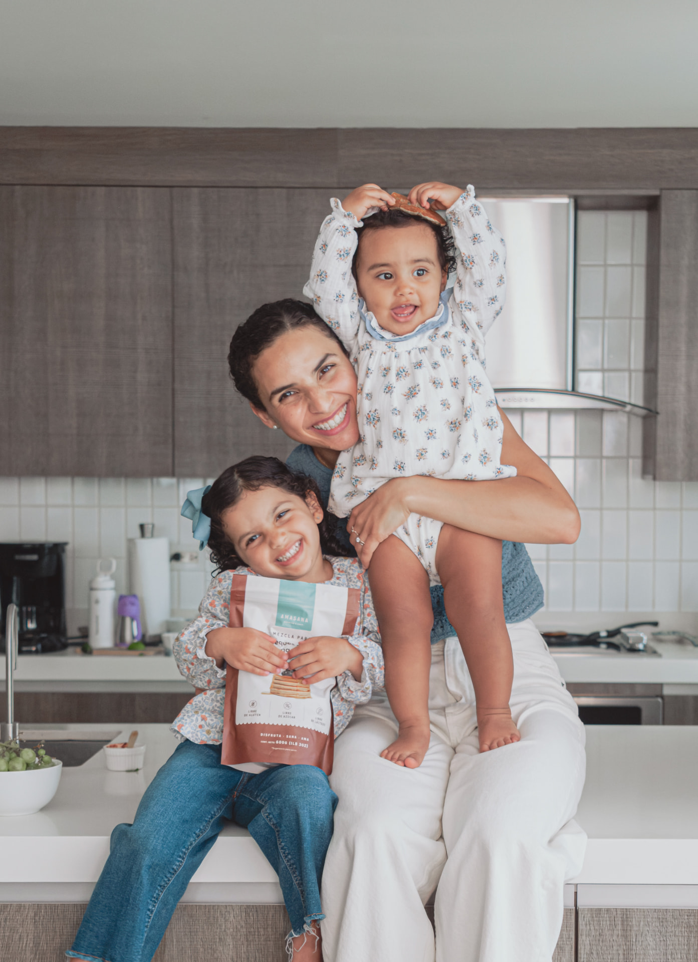 Victoria sonríe con sus dos hijas en la cocina, una de ellas sostiene una bolsa de AMASANA