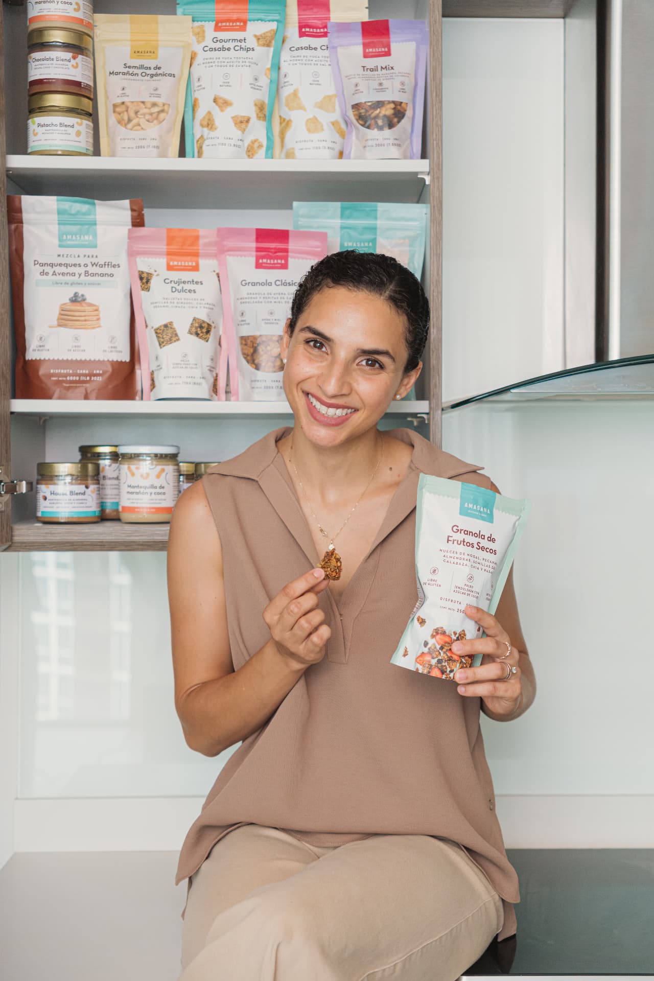 Victoria sonríe frente a la repisa de productos AMASANA con una bolsa de Granola de Frutos Secos